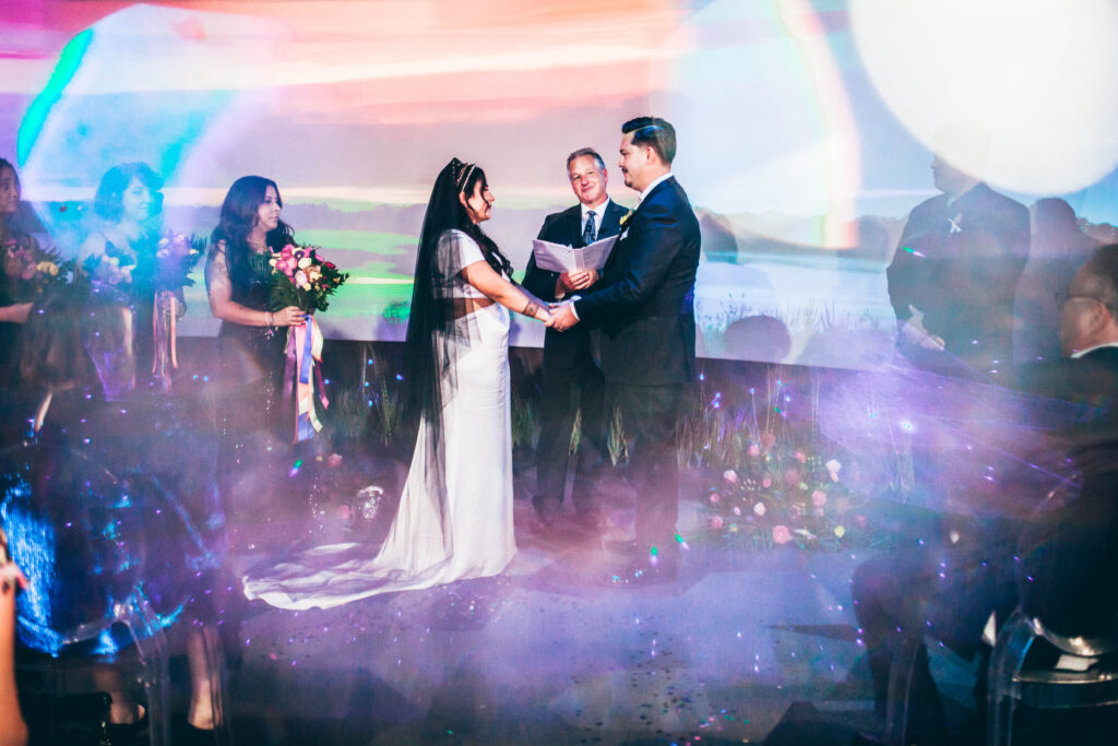 a wedding ceremony - bride in white dress and black veil to the left, with 3 bridesmaids in black sequin gowns behind her. Groom in  black suit to the right, with one groomsman in a black suit behind him. Officiant between the bride and groom in front of a large screen featuring Florida everglades.