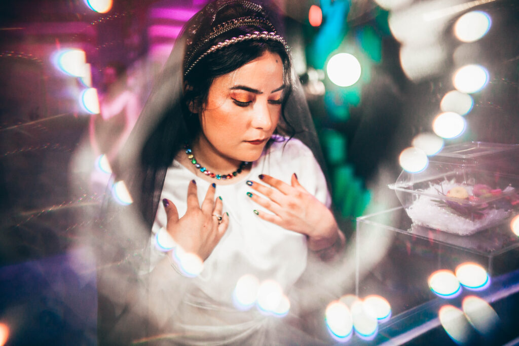 a bride's face looking down wearing a rainbow gem necklace, a gold beaded headband, a rainbow gem headband, and a black veil