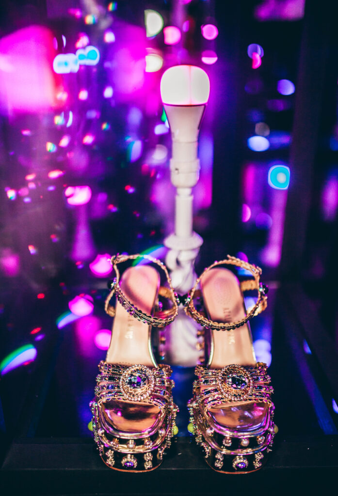 gold, rainbow gemmed platform heels on a black table in front of a pink light