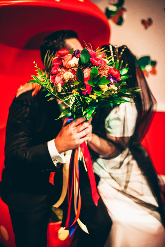 a bride and groom  sitting in a red chair hiding behind a bouquet of colorful flowers presumably kissing
