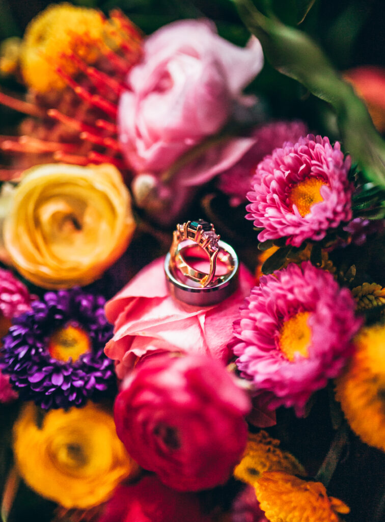 close up of 3 rings sitting on pink, purple, orange, and yellow flowers. One ring with a black center gem with two diamonds on each side of the center. One ring laying, a women's gold band. One ring laying, a man's silver band