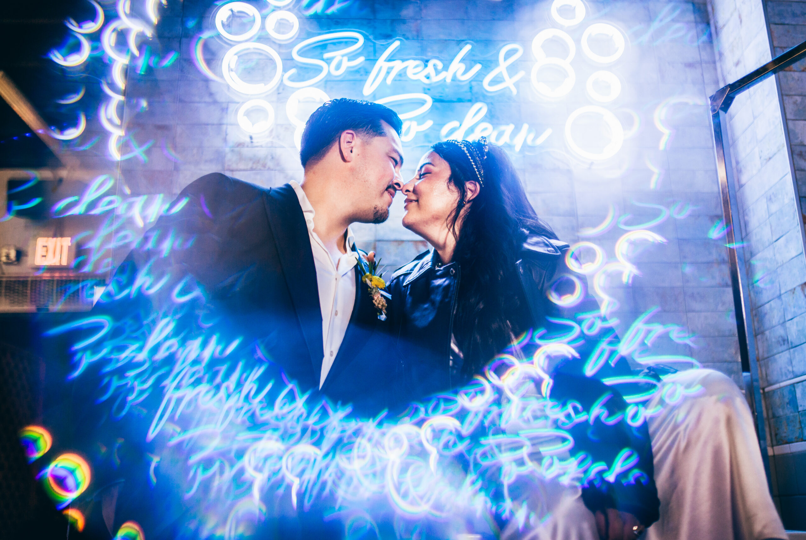a groom in a suit jacket and bride in a leather jacket and white dress facing each other surrounded by blue neon lights from a sign that reads "so fresh & clean"