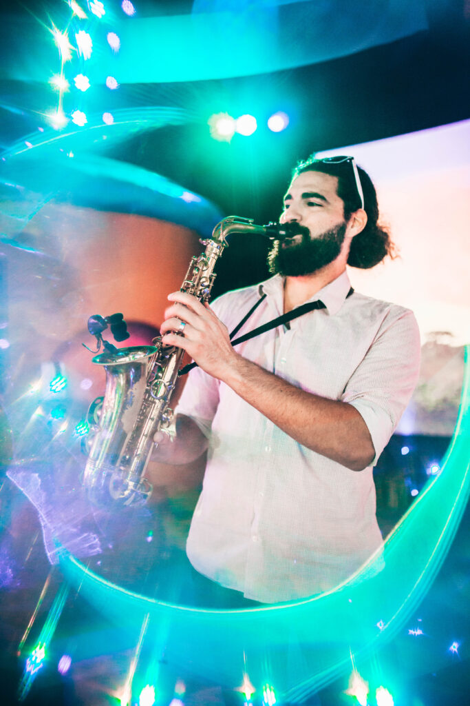 a bearded man in a white shirt, sunglasses on his head playing a saxophone surrounded by blue neon lighting