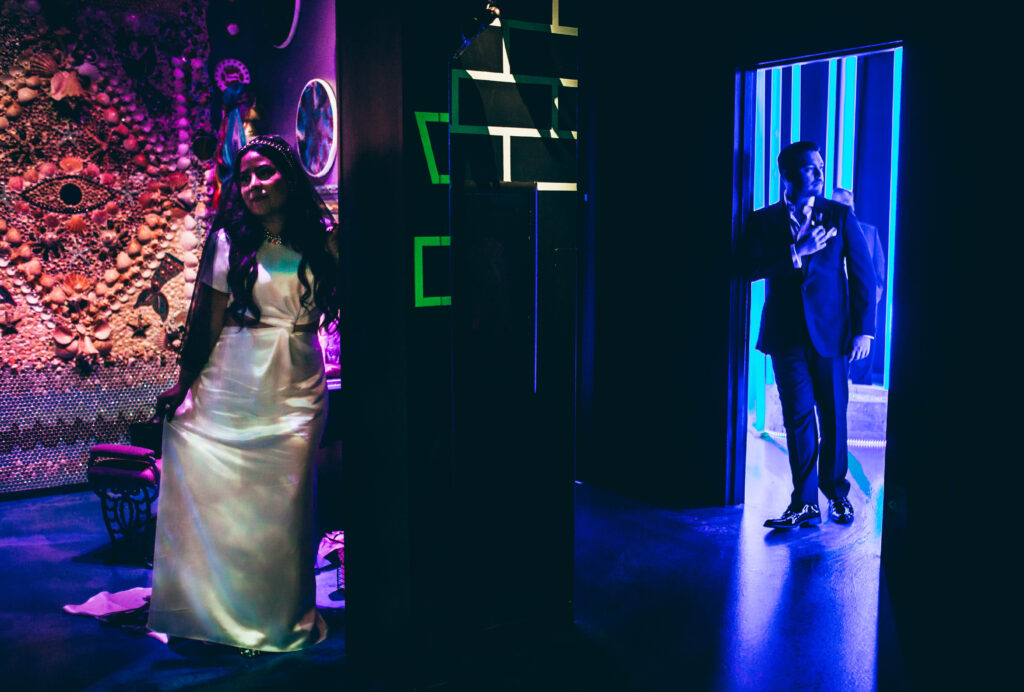 A bride in a white satin dress in a room with a wall covered in seashells arranged in an eye shaped pattern. Facing away from a groom in a doorframe of a room light with blue neon lights. They cannot see each other.