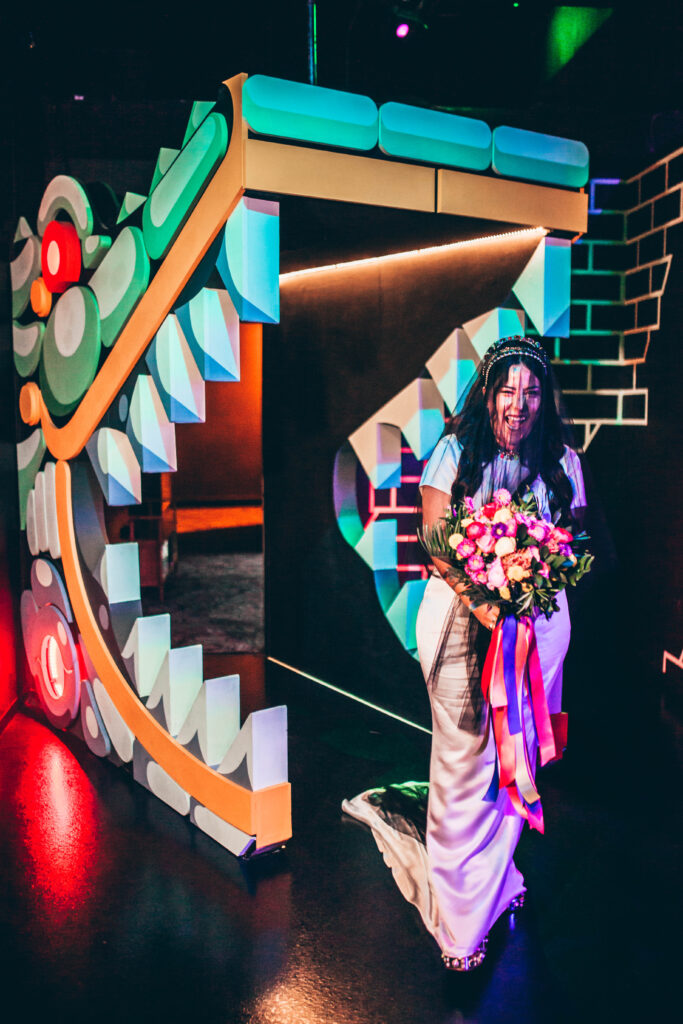 a bride with an excited expression wearing a black veil over her face, carrying a colorful bouquet of flowers draped with colorful ribbon, walking through the mouth of a large dragon art piece