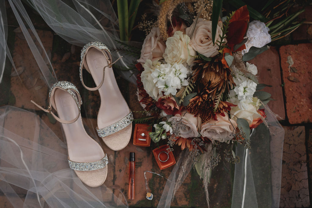 Flat lay arrangement of bejeweled open toe shoes, a man's wedding band, a bride's  diamond engagement ring and wedding band, a bouquet of flowers