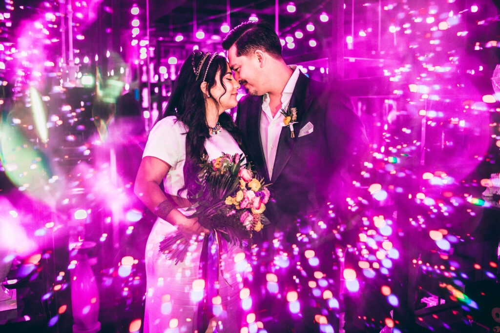 bride holding a colorful bouquet of flowers & a groom facing each other surrounded by pink neon lights