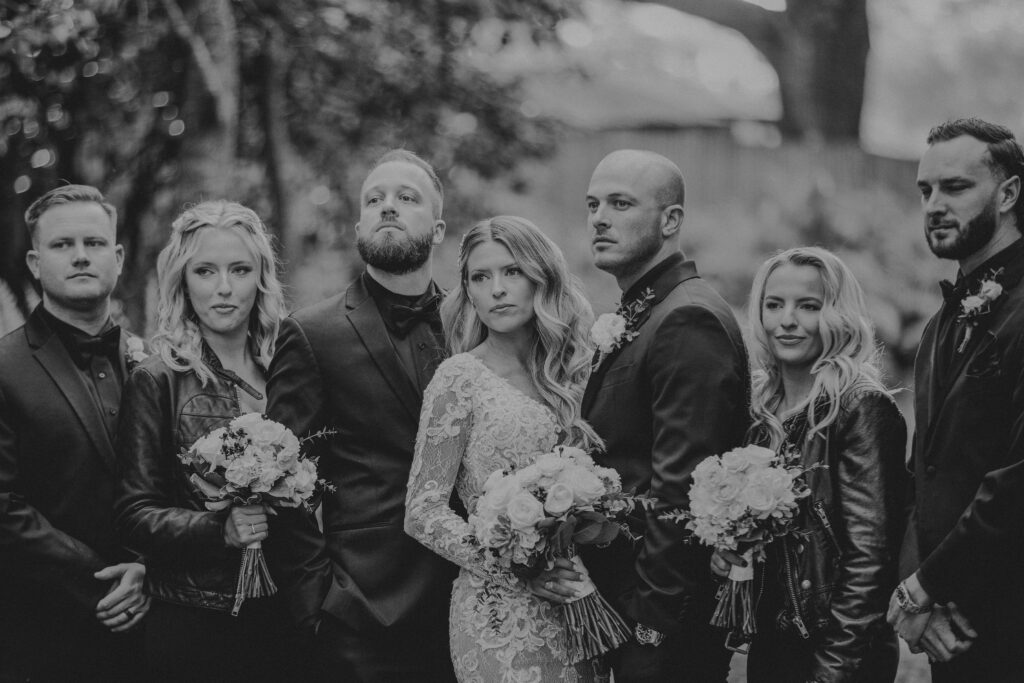 A bride, groom, two bridesmaids, and three groomsmen pose in a line with stoic facial expressions in a black & white photo