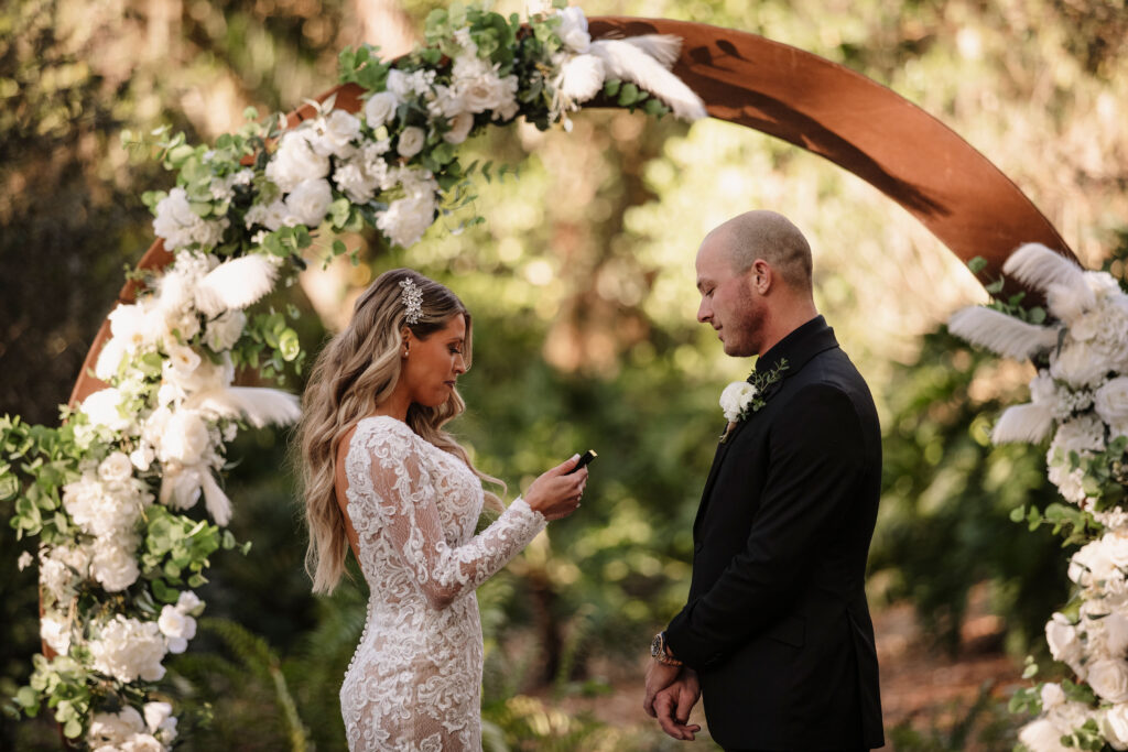 A bride in a white lace gown reading her vows to her groom in a black tuxedo standing in front of a circular backdrop covered in white flowers