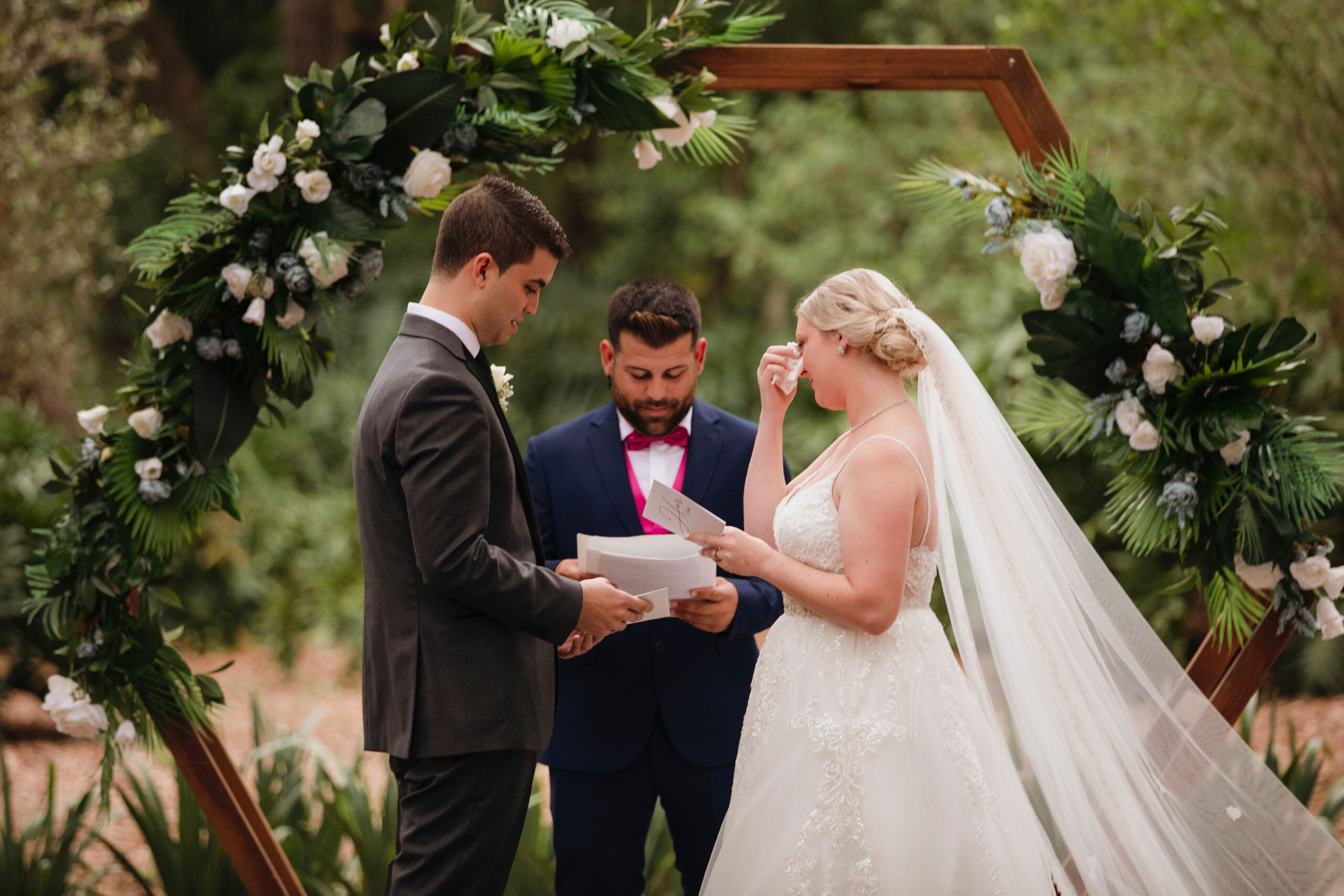 a bride, groom, and officiant stand in front of a six sided ceremony backdrop. Tropical greenery and white flowers on the left and right side of the backdrop. The bride is wiping a tear away with a tissue
