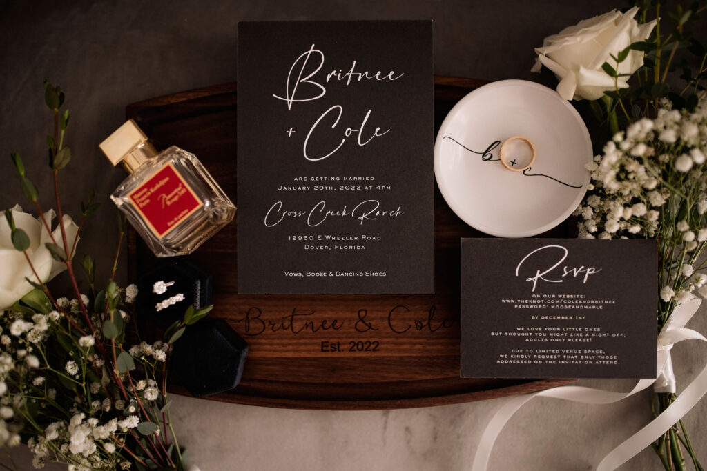 a flat lay arrangement of a black wedding invitation with white script, a small white bowl with a gold ring inside, a bottle of perfume, and a black velvet ring box  with 2 diamond rings surrounded by white flowers