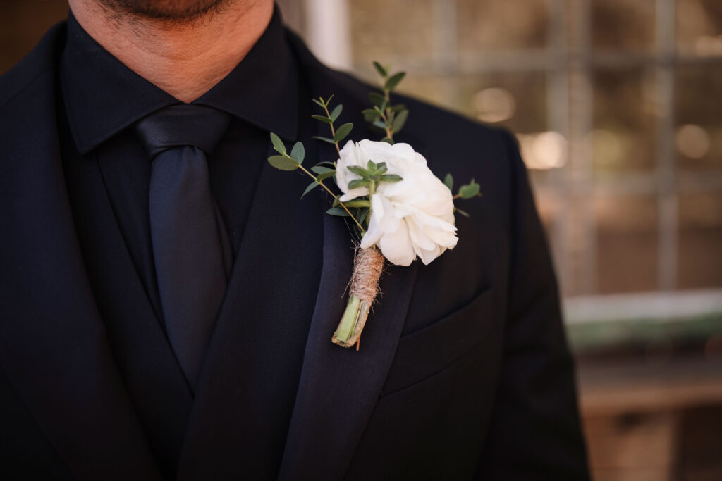 a groom's details - black tuxedo jacket, bllack vest,  black button down shirt, black euro tie and a white single flower pinned to his lapel