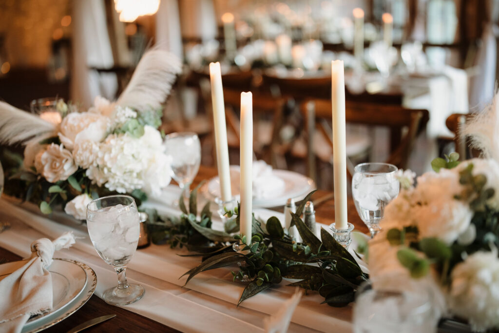 a wood farmhouse table dress with a flowy white table runner, greenery garland white flower arragements, white taper candles, and place settings