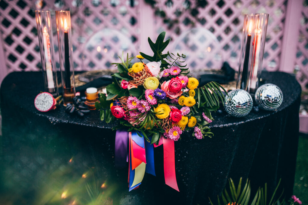a warm colored bouquet of pink, yellow, and purple mums, orange proteas, and greenery resting on  rainbow ribbons, and black sequin  linen sweetheart table
