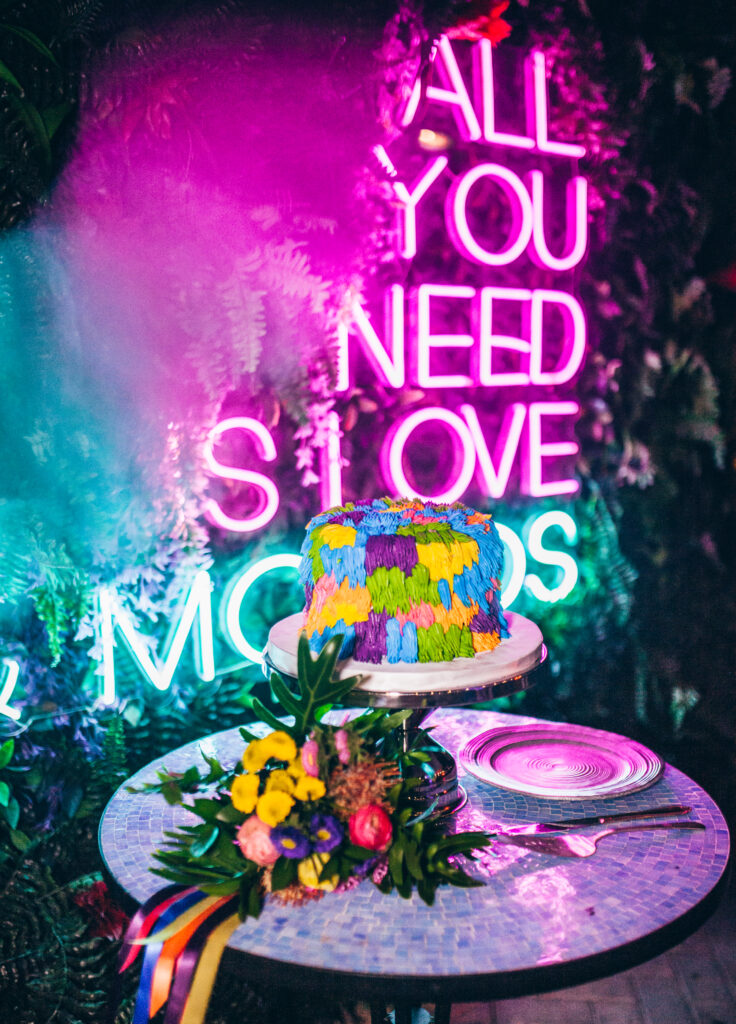 a single tier cake decorated in purple, yellow, pink, green, and blue shag frosting sitting on a mosaic bistro table next to a small colorful bouquet of flowers, a small plate, and a cake knife and server.
In front of a pink neon sign reading "ALL YOU NEED IS LOVE & MOJITOS"