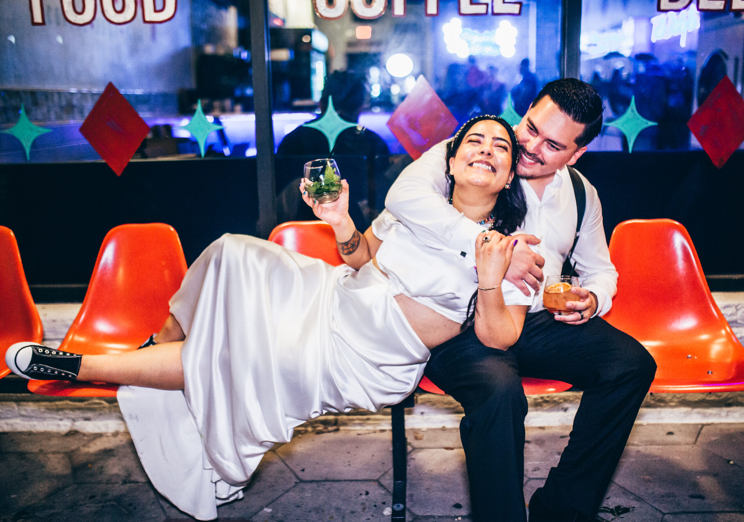 A bride laying across a row or orange bowling alley type seating, holding a stemless wine glass containing mint leaves, she is leaning into a groom
Groom sitting with his arm around the bride smiling, holding a stemless wine glass containing an orange slice and orange drink