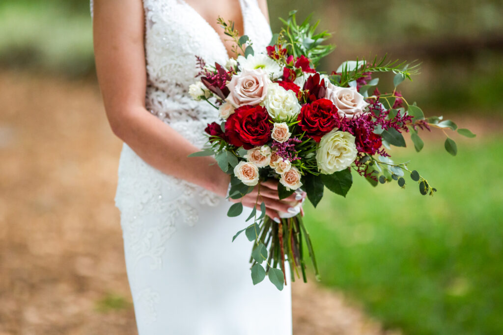 bride holding a colorful bouquet in shades of red and pink. red gardden roses, blush pink tea roses, ivory roses, and greenery