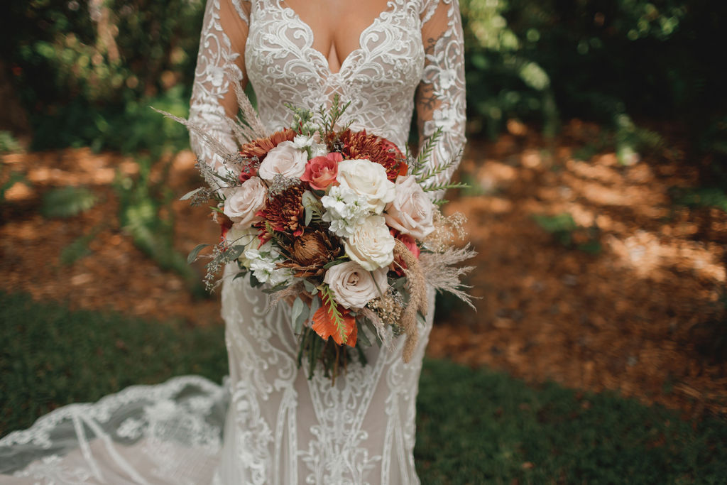 a bride holding a fall, boho style bouquet  of white and ivory roses, dark orange and golden chrysanthemums, orange leaves, brown protea, and golden wheat