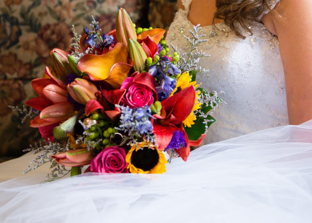 a rainbow color bouquet in jewel tones. red lilies, dark pink roses, orange calla lilies, green berries, yellow sunflowers, and blue filler flowers